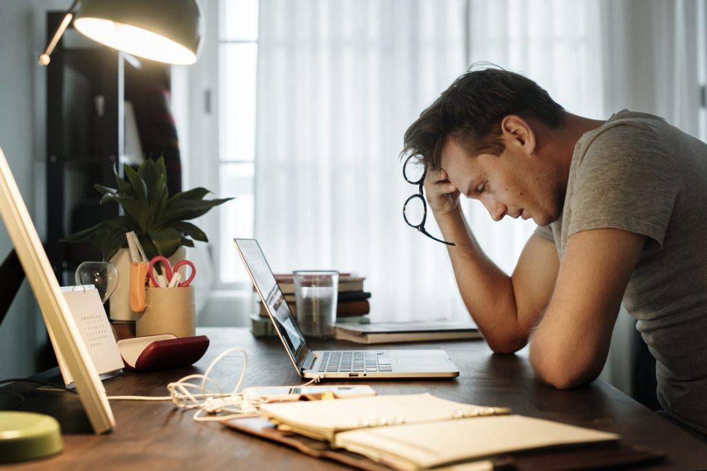 Man at desk near computer, holding glasses and looking stressed.
